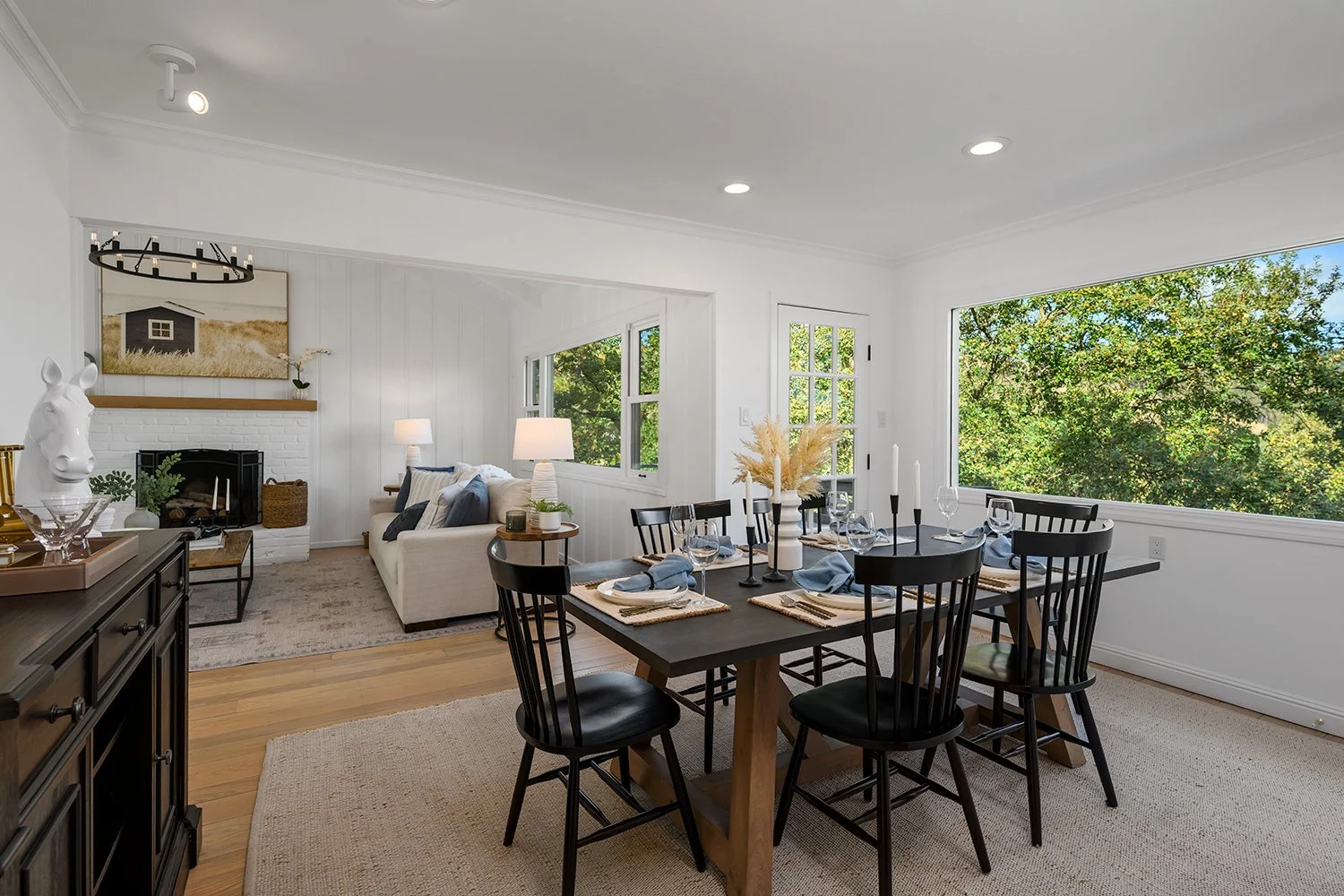 Bright dining and living room with large windows, black dining table set for dinner, white sofa, fireplace, and modern decor.