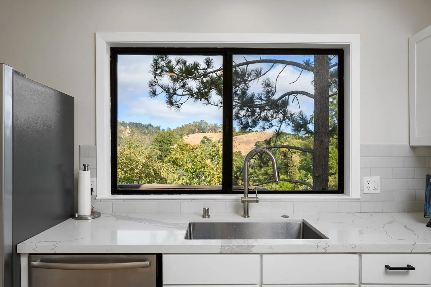 Modern kitchen with white countertop, stainless steel sink, window showing trees and hills outside.