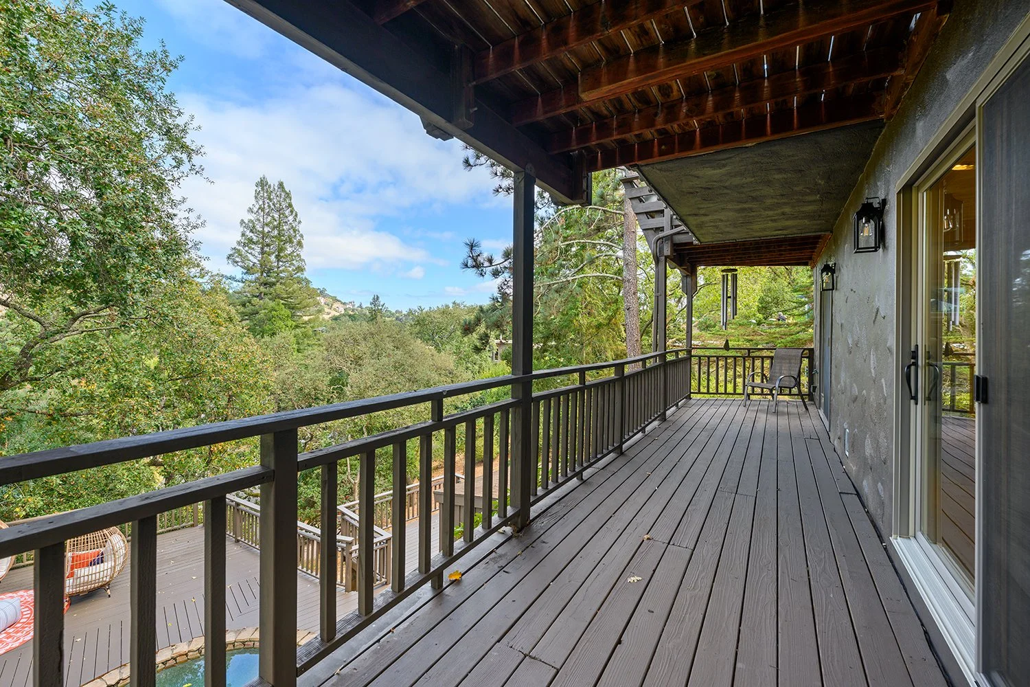 Wooden balcony with black railing, outdoor chairs, and a view of green trees and a backyard with a pool.