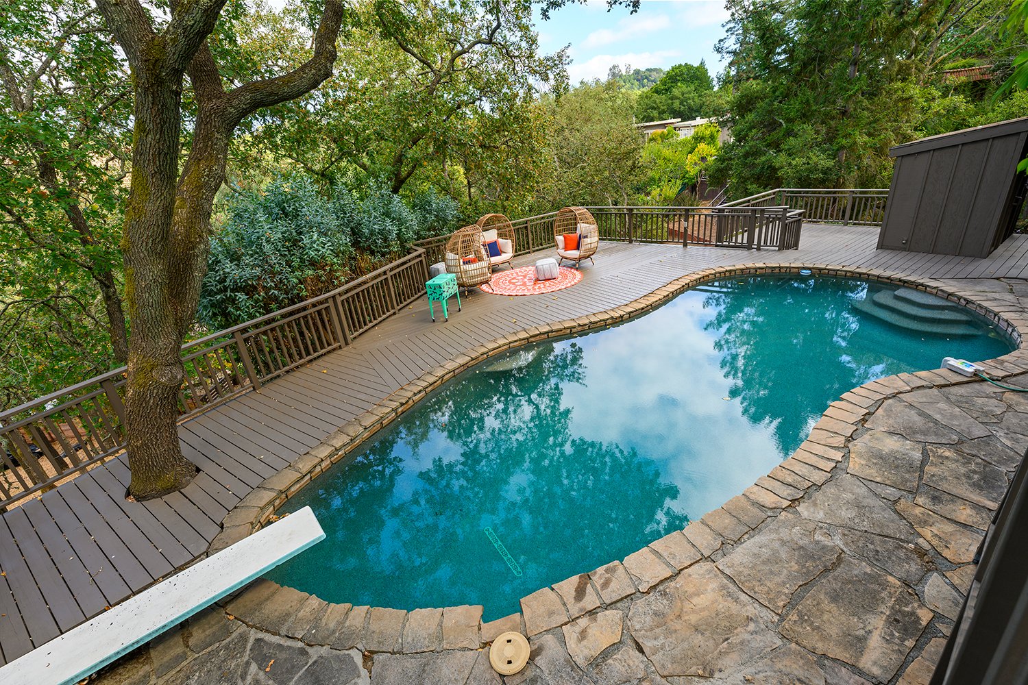 A backyard with a swimming pool surrounded by wooden decking, greenery, and trees. There is an outdoor seating area with wicker chairs, colorful cushions, a small rug, and a side table.