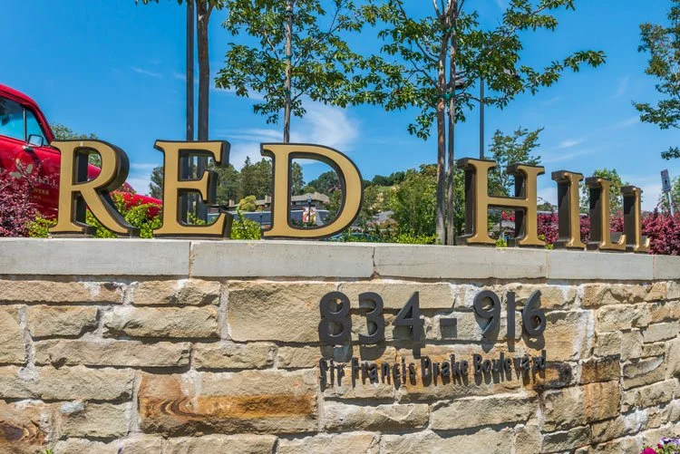 Large gold letters spell out 'RED HILL' on a stone wall, with a street address and street name in black below, in front of trees and a clear blue sky.