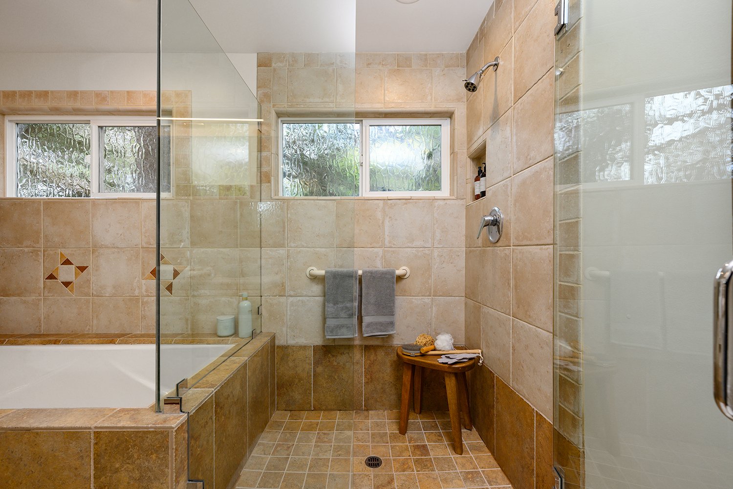 Modern walk-in shower with beige tiles, a bathtub, a small wooden stool with towels and toiletries, frosted windows, and a glass partition.