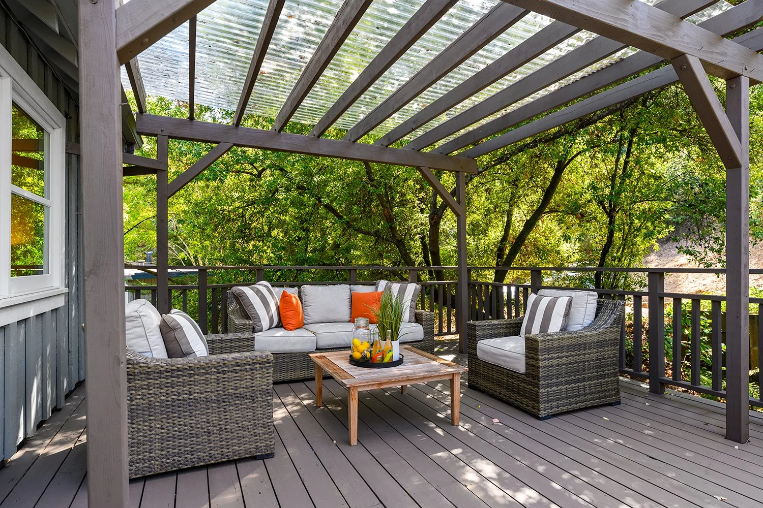 Outdoor deck area with wicker chairs and a sofa, a wooden coffee table with a tray of drinks and a plant, surrounded by green trees and a wooden railing.