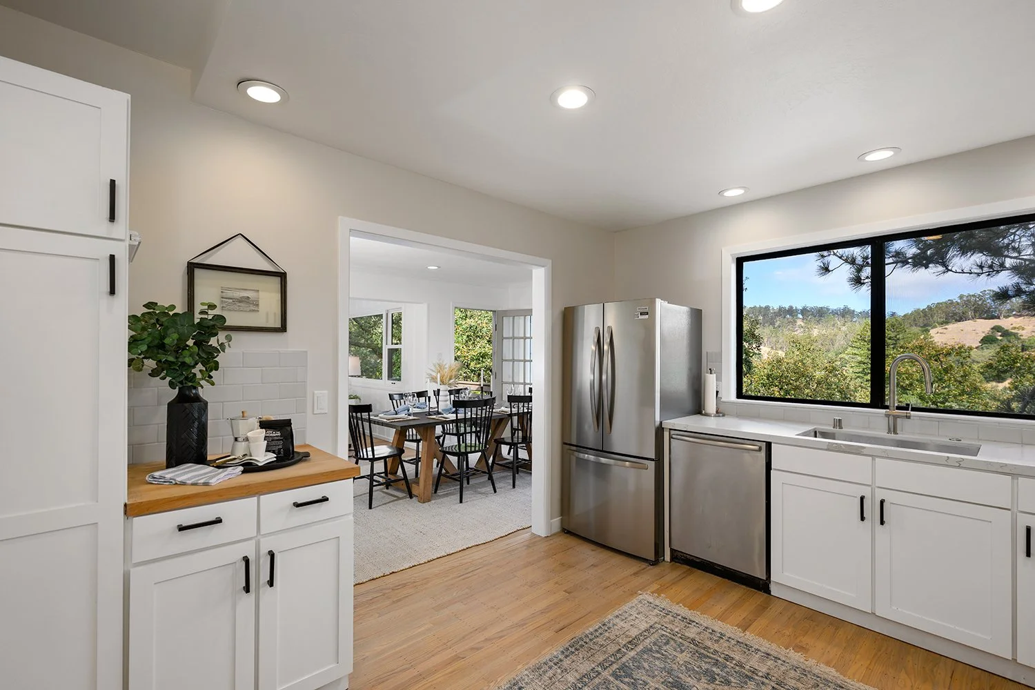 Modern kitchen with white cabinets, stainless steel refrigerator, large window with view of trees and hills, adjacent dining area with black chairs and wooden table.