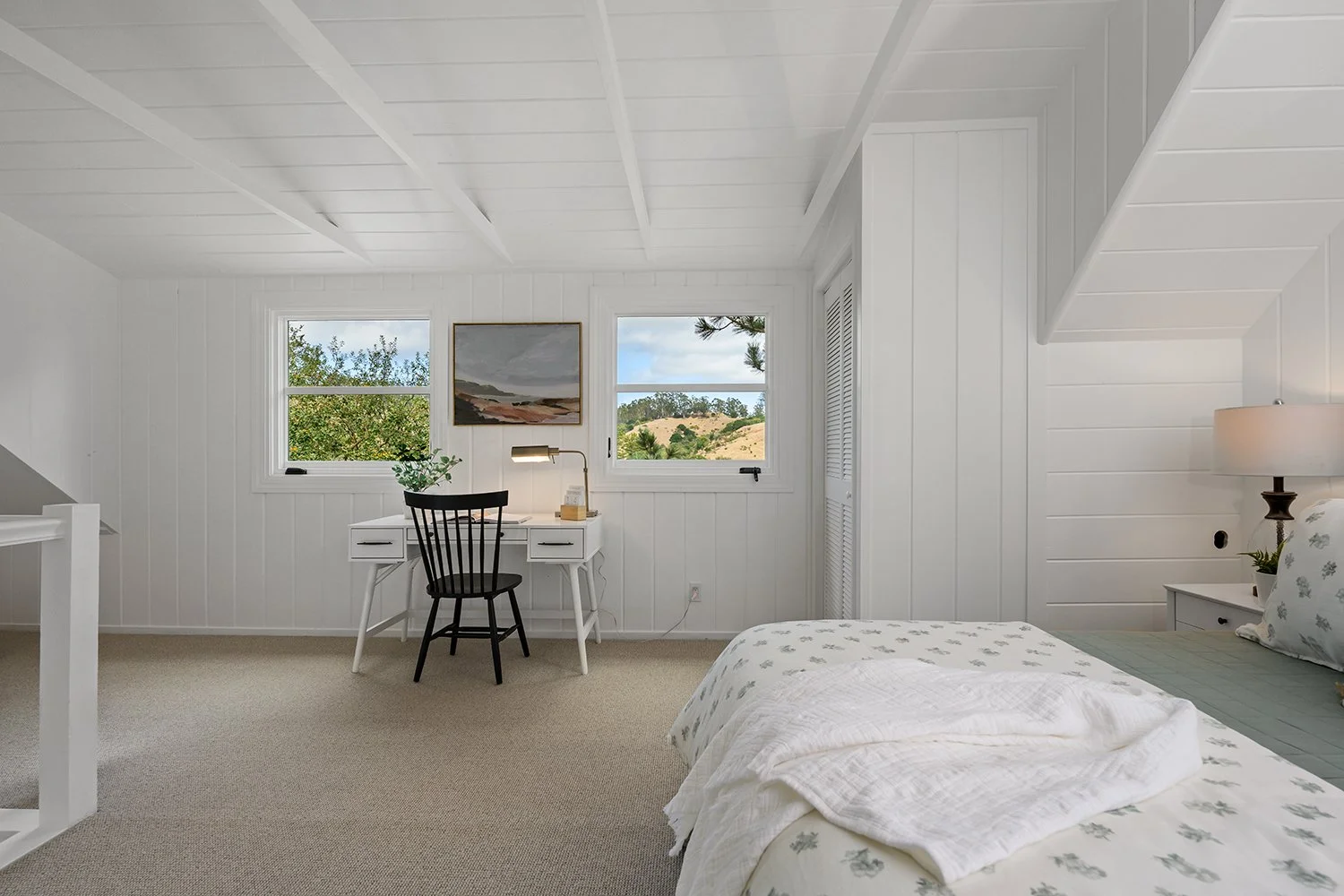 A bright, minimalist bedroom with white shiplap walls, a bed with floral bedding, a white bedside table with a lamp and plant, a small white desk with a black chair, and two windows showing outdoor greenery and hills.