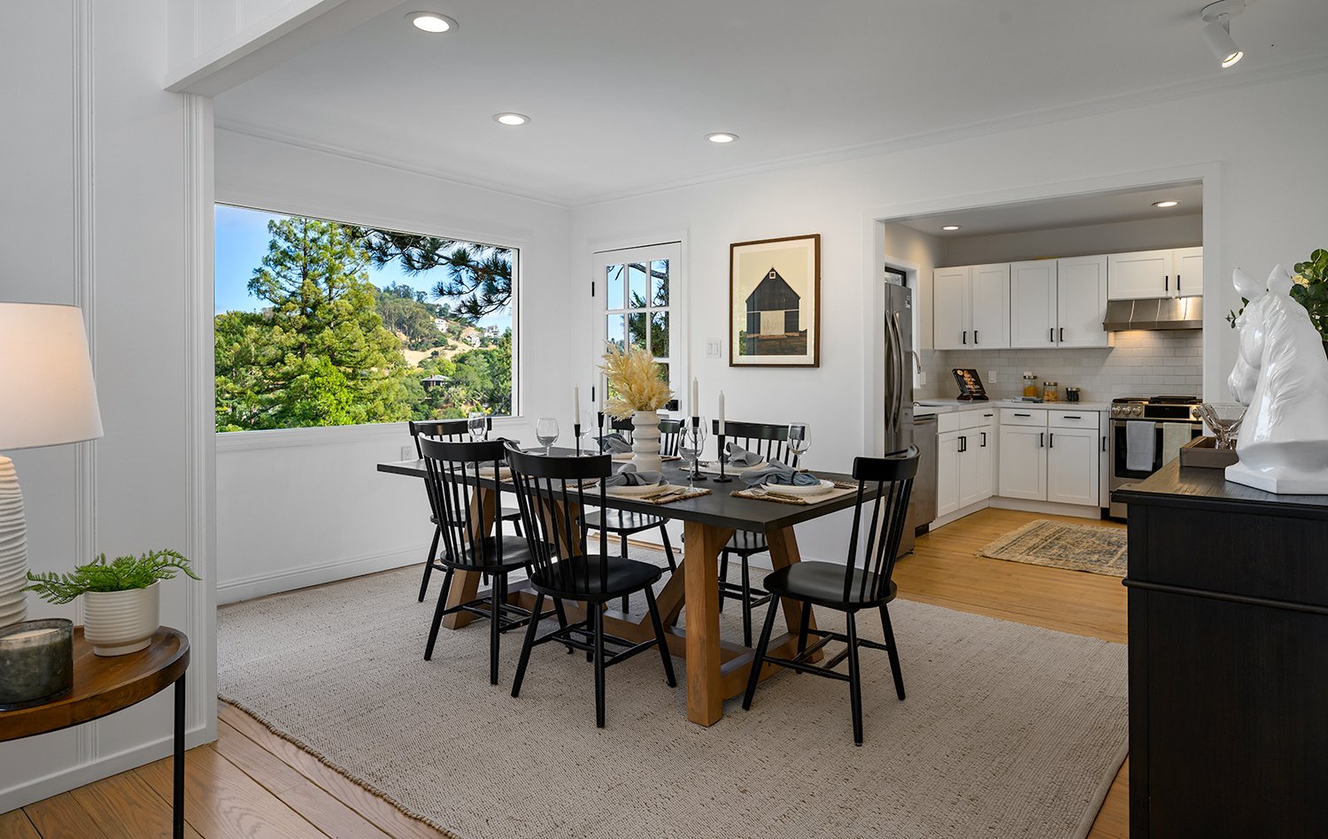 Dining area with black table and chairs, set with gray napkins, glassware, and a white vase with dried flowers, adjacent to a white kitchen with stainless steel appliances and a large window showing a green outdoor view.