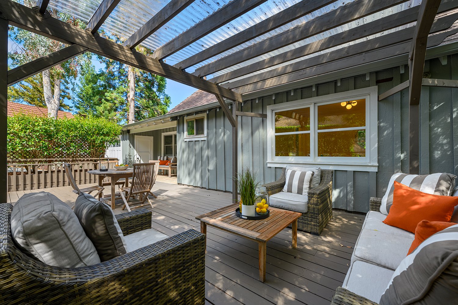 Outdoor patio with wicker sofas, orange and striped cushions, wooden coffee table with plants and lemons, dining table with chairs, shaded by a pergola, with a green yard and trees in the background.