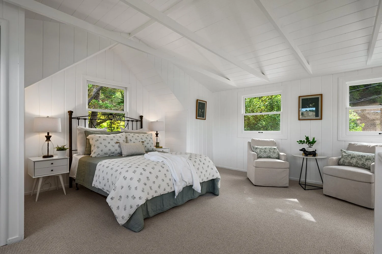 A bright, cozy bedroom with a white-paneled ceiling and walls, a black metal bed with white and floral bedding, two nightstands with lamps, and two armchairs with side table and decorative pillows. Multiple windows reveal green trees outside.