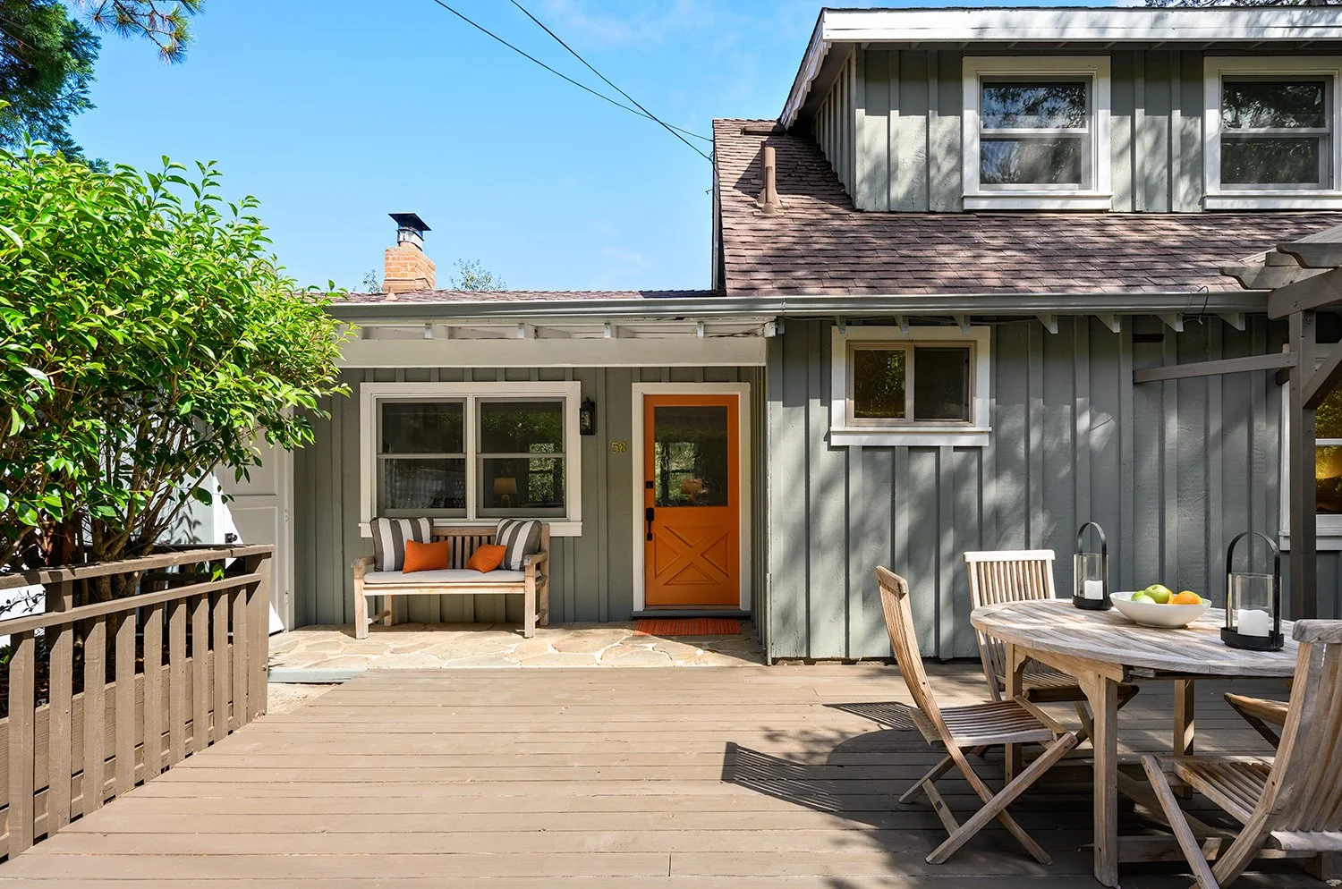 The image shows a backyard patio area with a wooden deck, a round wooden table with four chairs, a bowl of fruit, and lanterns. There is a small bench with striped pillows beside a window, and a door with an orange frame leading into a house. The hou
