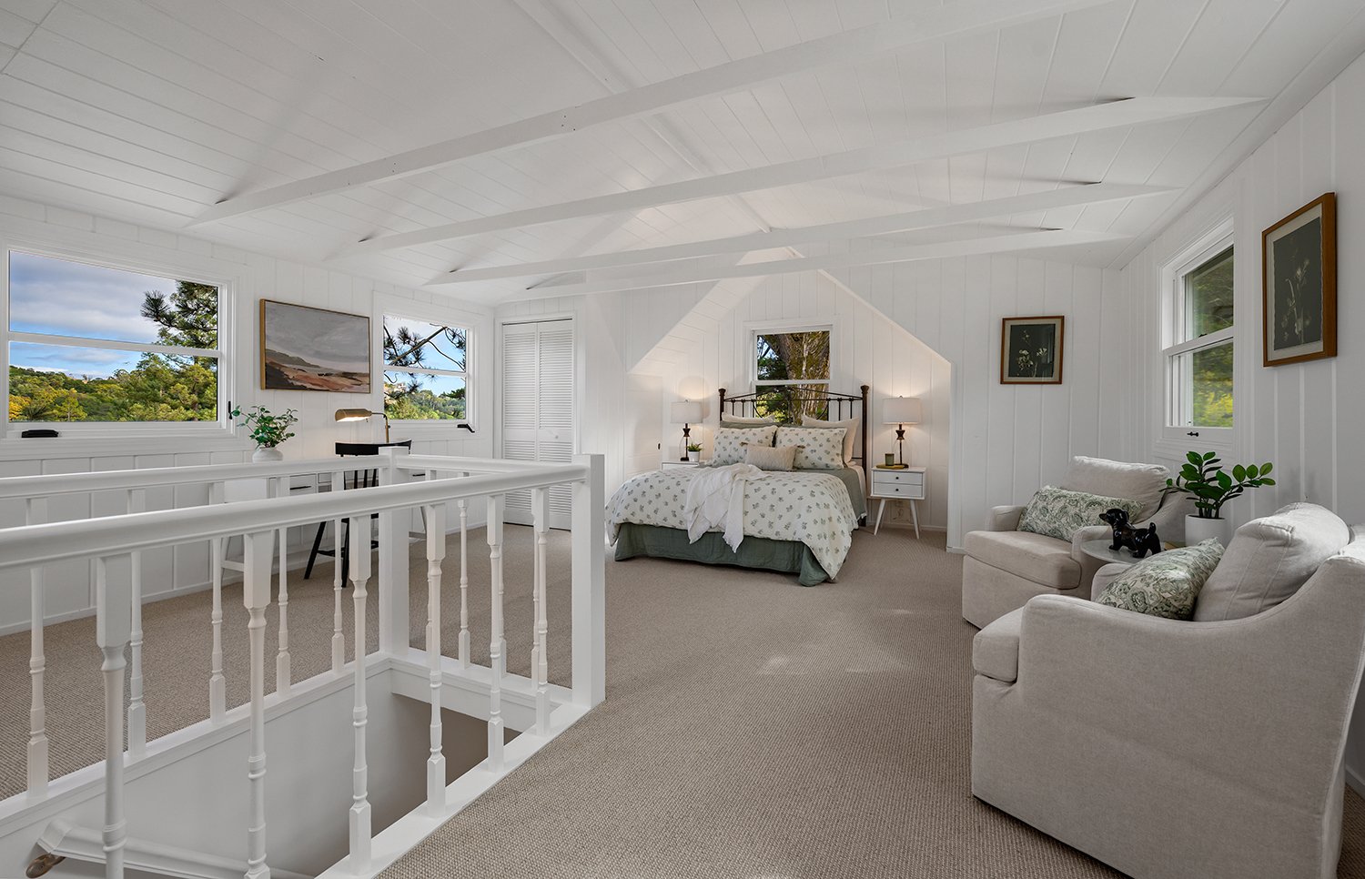Bright white bedroom with sloped ceilings, large windows showing green landscape, a bed with patterned bedding, two nightstands with lamps, a sitting area with two chairs, framed artwork, and minimal decor.