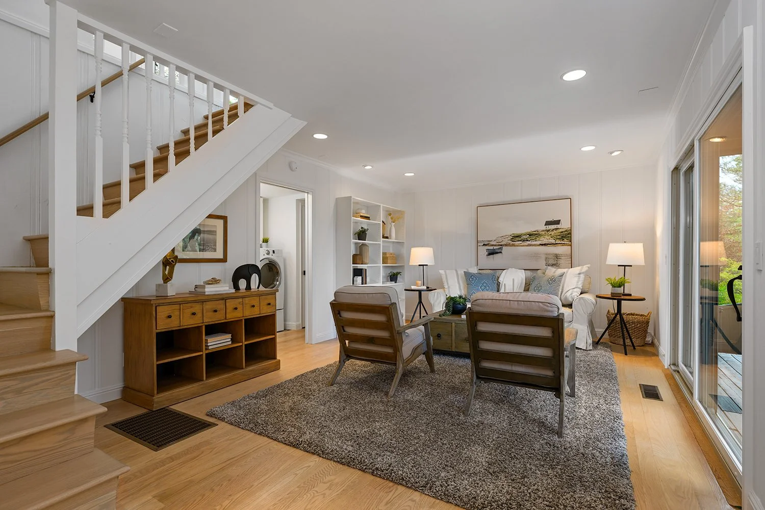 Living room with white walls, hardwood floors, a staircase to the left, a sitting area with a white sofa, wooden chairs, and a coffee table, side tables with lamps, and a large painting on the wall. There is also a sliding glass door leading outside.