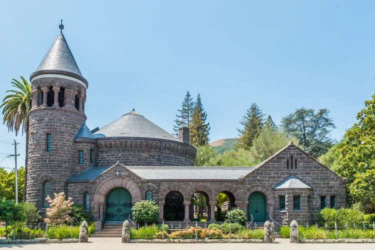A stone castle-like church with a tall tower, arched entrance, surrounded by green trees and plants, under a clear blue sky.