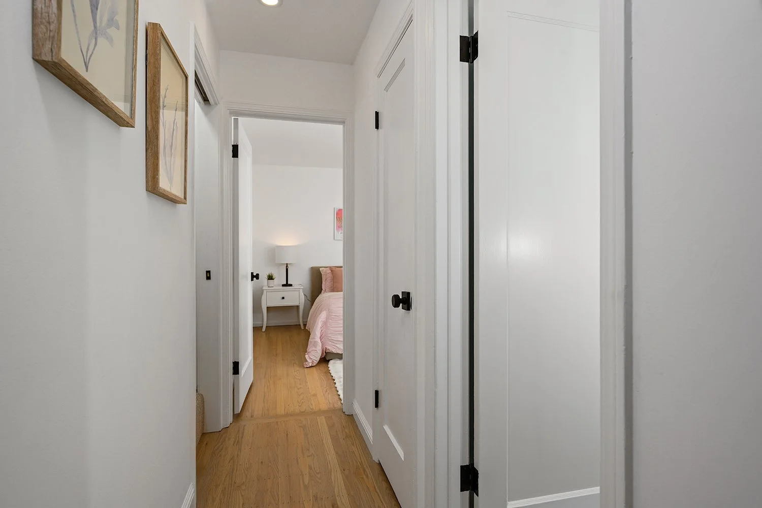 A hallway leading into a bedroom with a pink bedspread, a white nightstand, and a lamp.