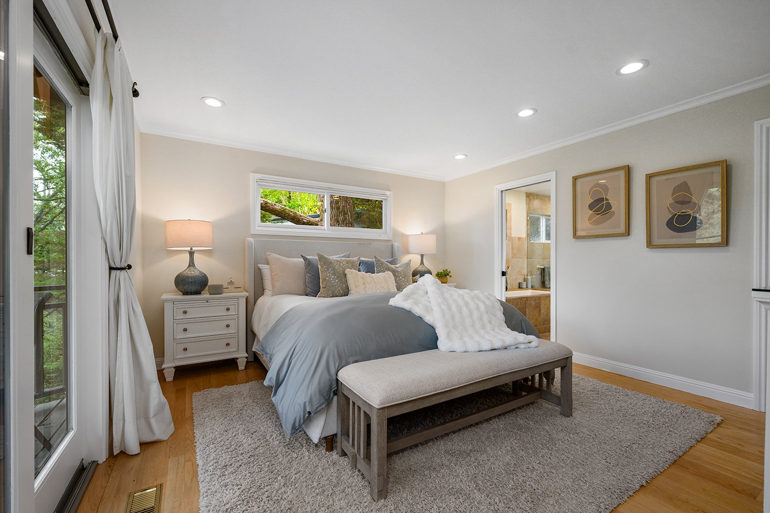A cozy bedroom with a bed, multiple pillows, nightstands with lamps, a bench at the foot of the bed, a window overlooking trees, hardwood flooring, and a bathroom visible through an open door.