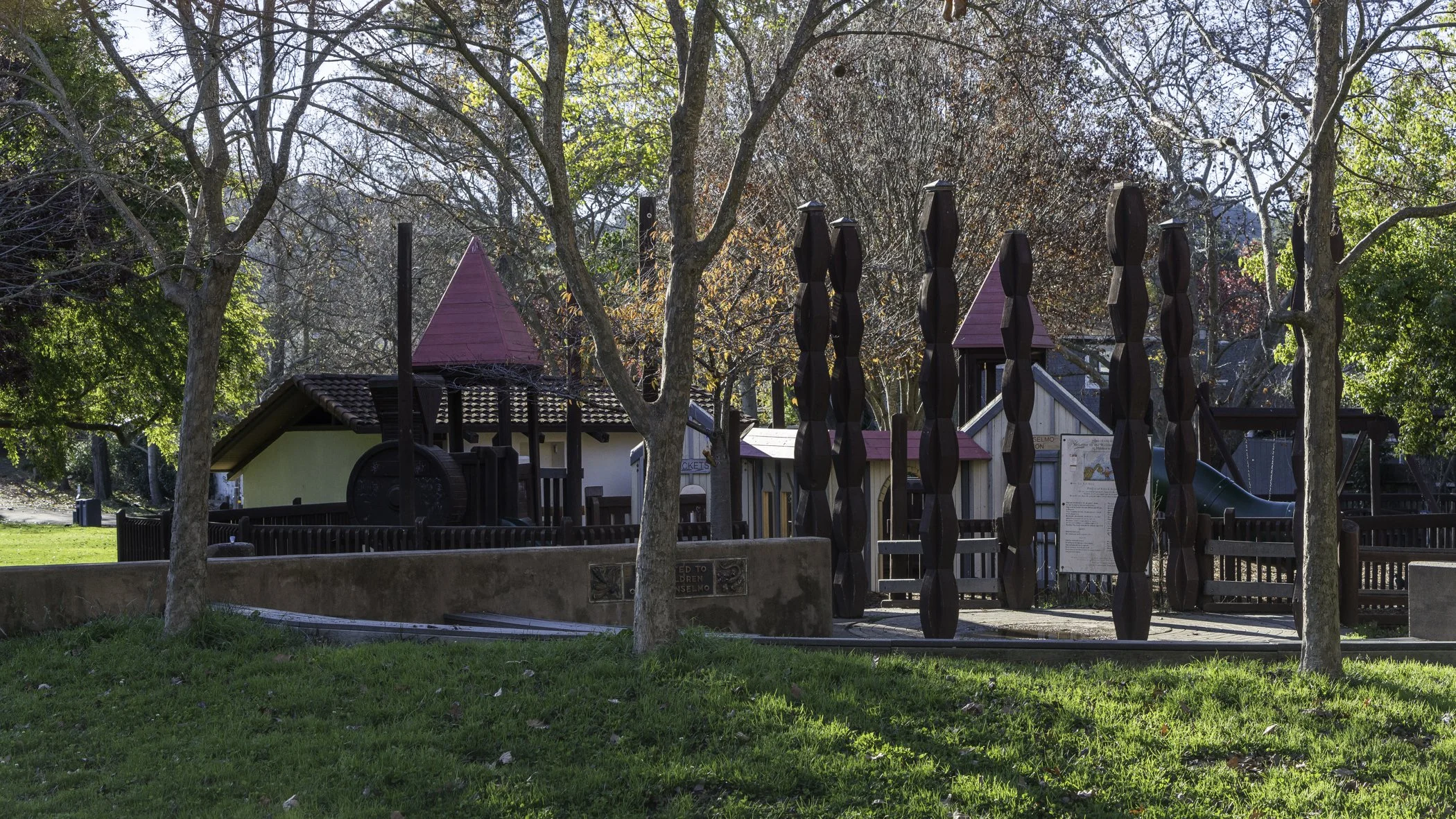 A playground surrounded by trees with autumn foliage, featuring play structures, slides, and a fenced area.