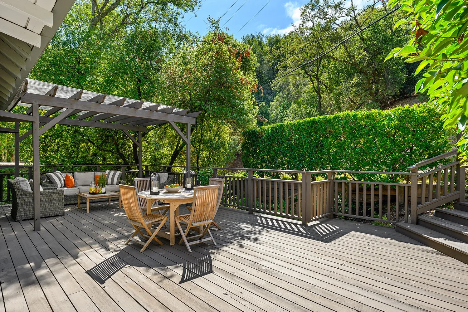 A wooden outdoor deck with a round dining table and four chairs. To the side, there is a seating area under a pergola with a sofa and armchair. The deck is surrounded by lush green trees and bushes, with blue sky and clouds visible in the background.