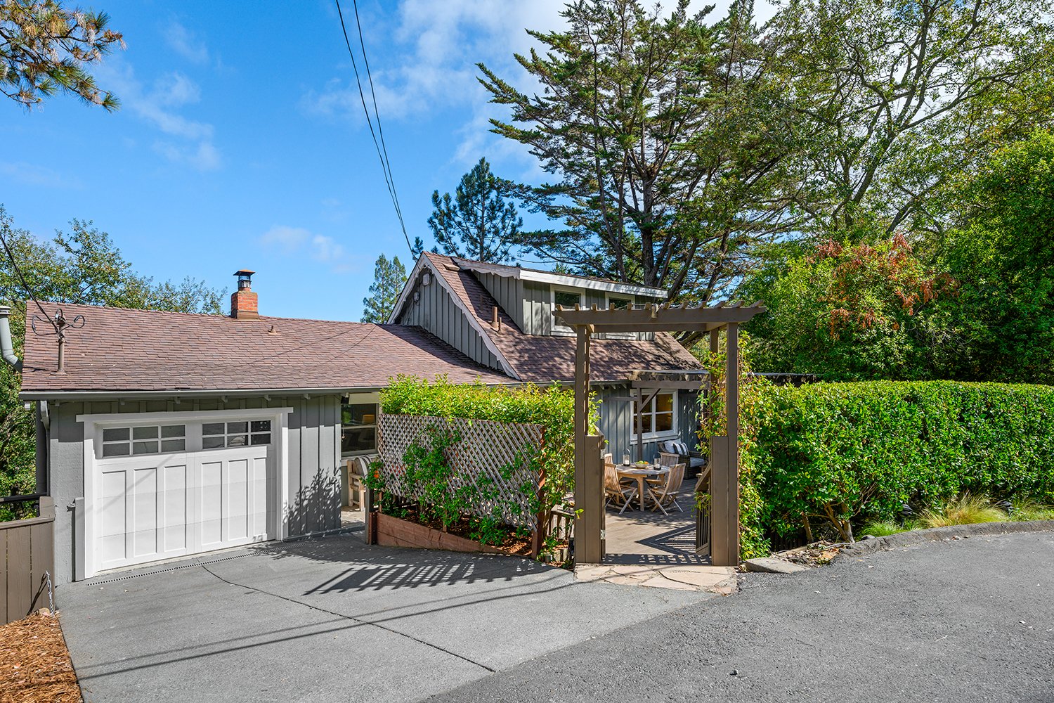 A house with a garage, a wooden pergola, outdoor dining furniture, lush green hedge, and tall trees under a blue sky.