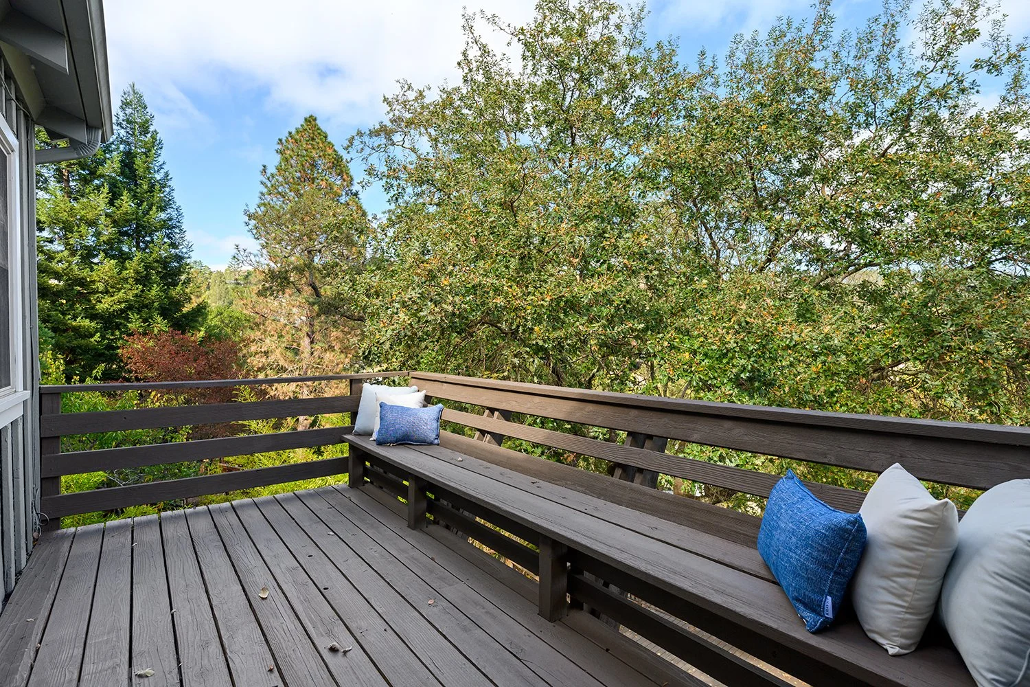 Wooden balcony with a built-in bench decorated with white and blue pillows, overlooking a green forest with tall trees under a partly cloudy sky.
