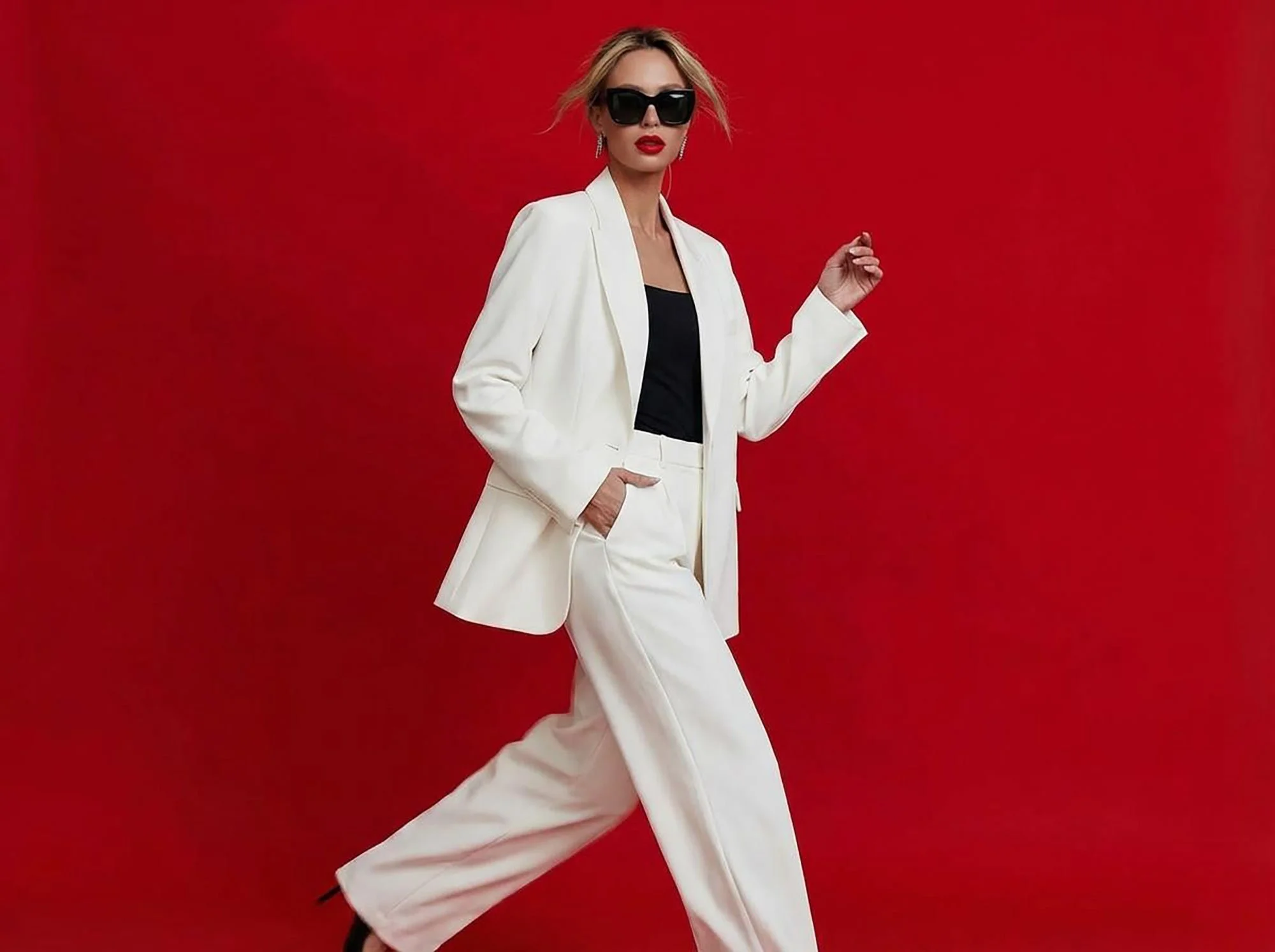 A woman wearing a white suit with a black top, sunglasses, and red lipstick standing against a red background.