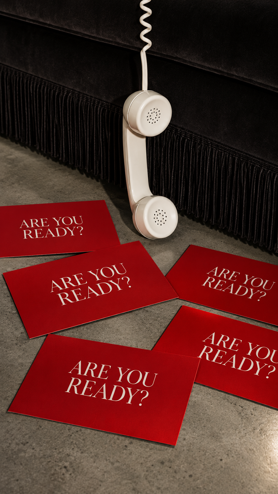 A vintage white telephone hangs against a black sofa with fringed skirt. On the floor, six red cards with white text read 'Are You Ready?' in different orientations.