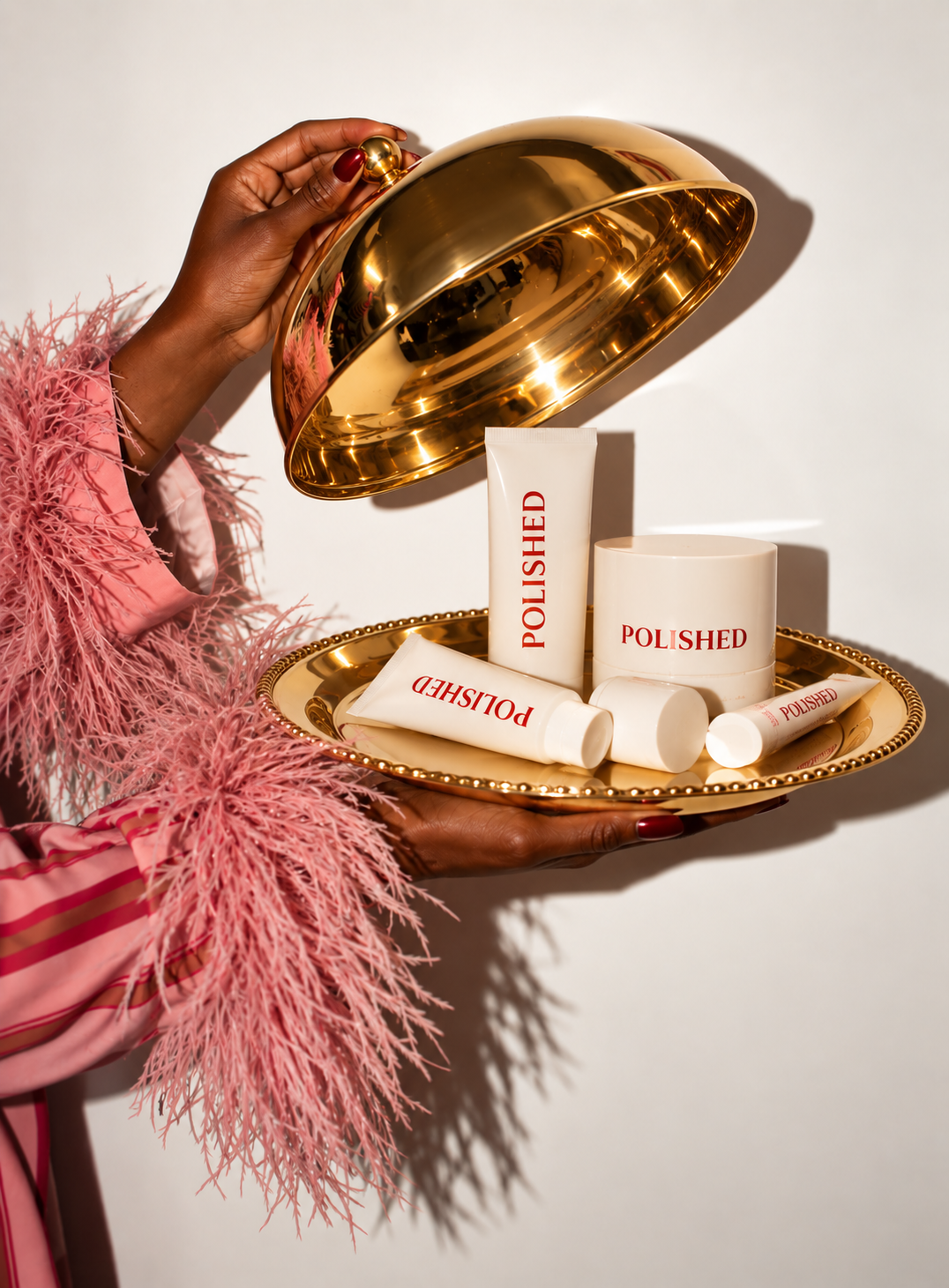 A person holding a gold tray with a mirrored lid, revealing skincare products labeled 'POLISHED', including tubes and a jar, on a white background.