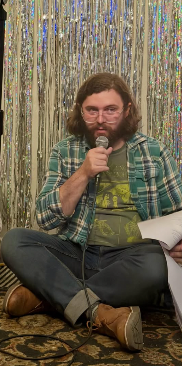A man with glasses and long hair, sitting cross-legged on a patterned rug, holding a microphone, and reading from papers, with a shiny foil curtain backdrop.