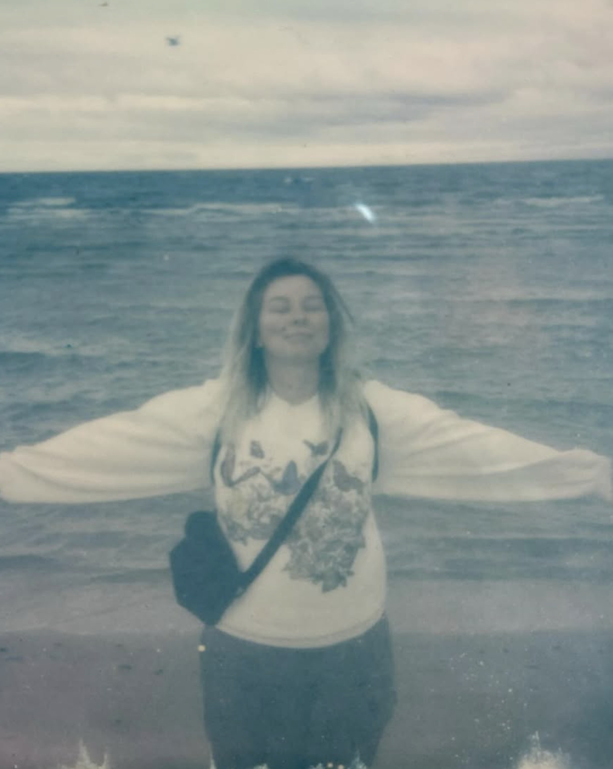 A woman standing in the ocean with her arms outstretched, wearing a white sweatshirt and a black crossbody bag, smiling with her eyes closed, waves in the background, overcast sky.