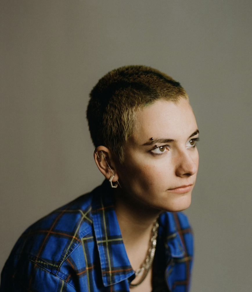 A young person with short hair and multiple facial piercings, wearing a blue plaid shirt, looking to the right.