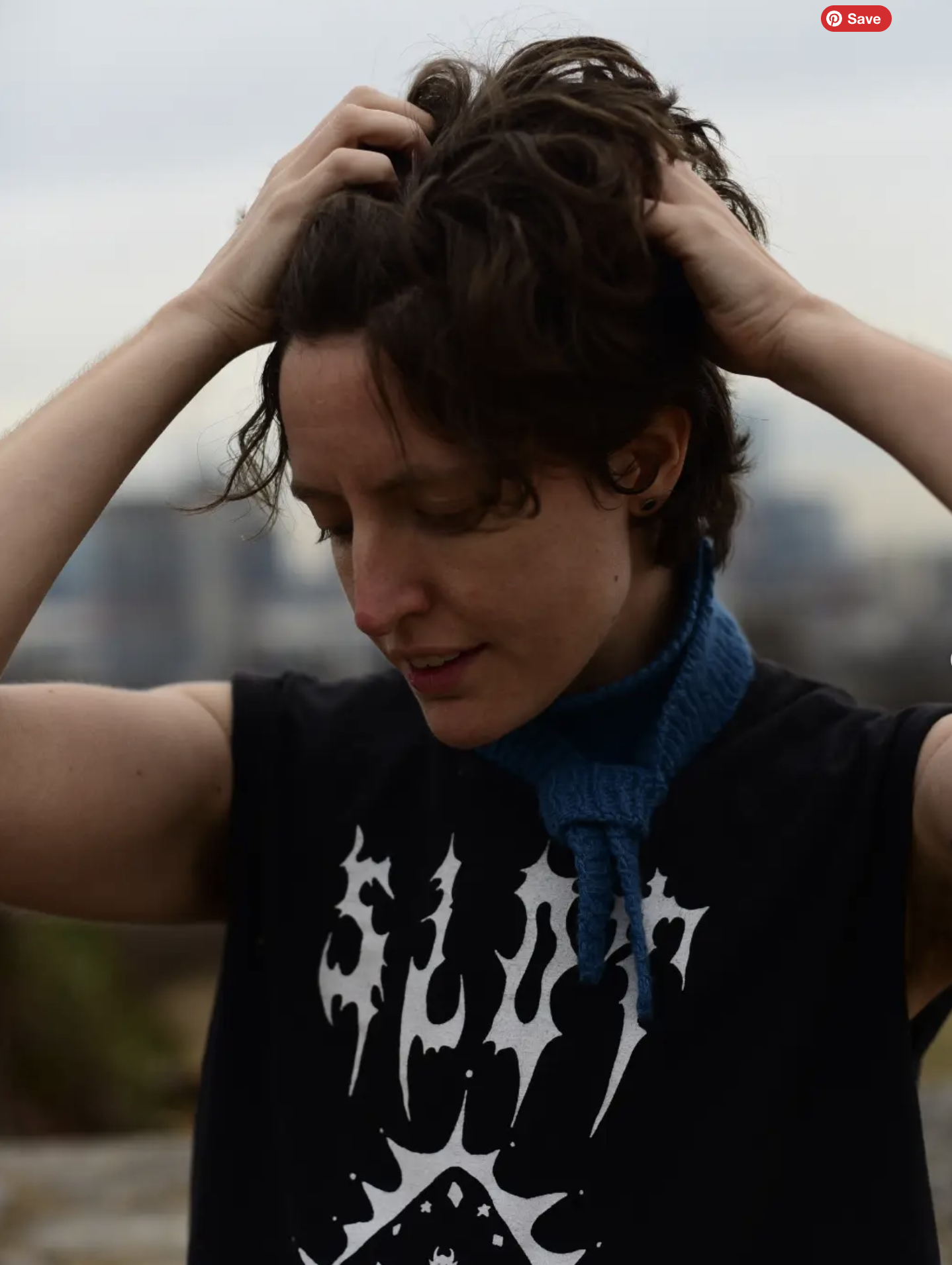 A person with short brown hair, wearing a black T-shirt with a white graphic design, and a blue neck gaiter, holding their head with both hands, smiling with eyes closed, outdoors.