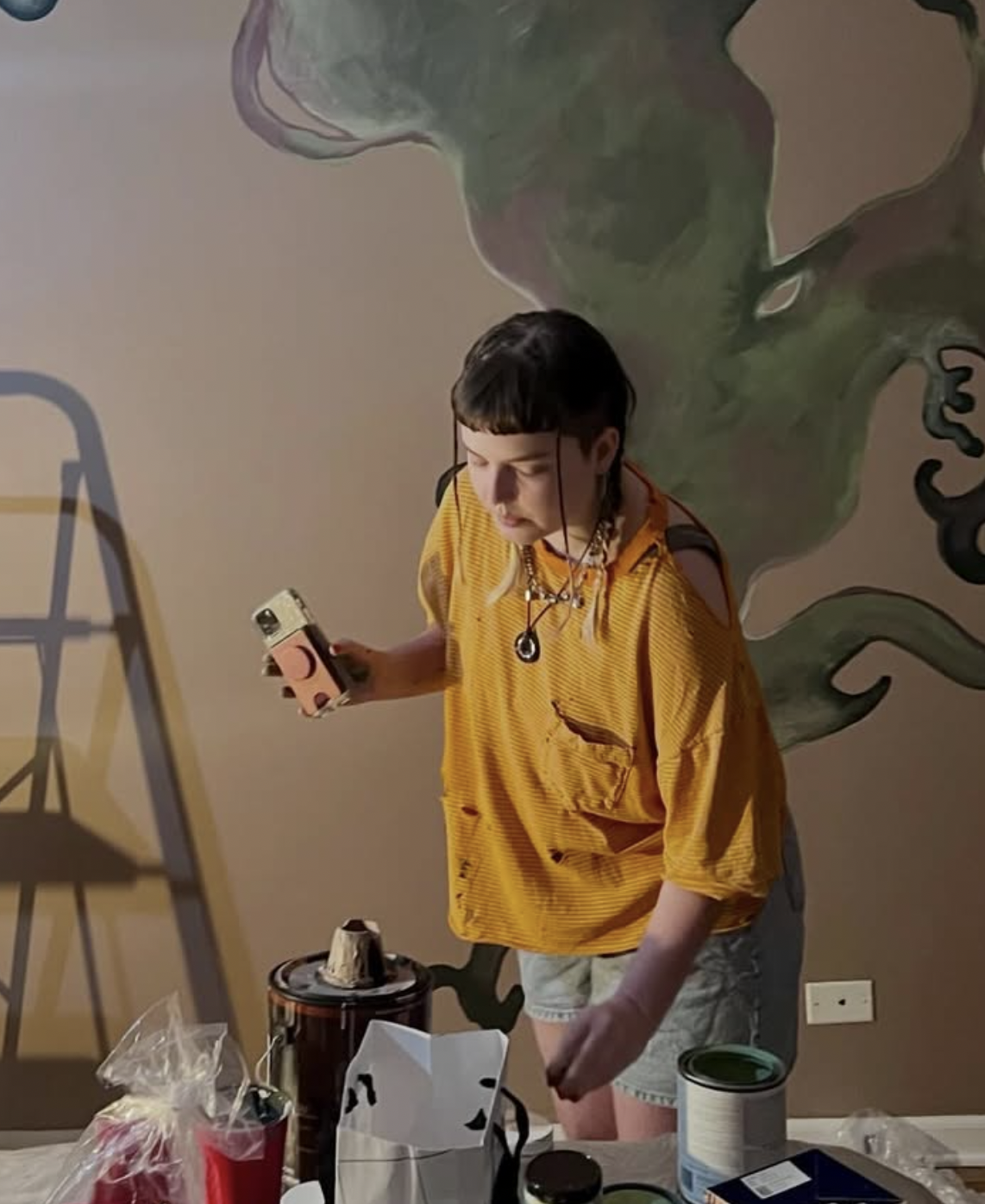 A young person with dark hair wearing a yellow shirt, shorts, and a necklace, standing at a table with various items, looking at a phone. They are in front of a colorful abstract mural on the wall.