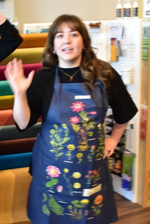 A young woman with brown hair, wearing a black top and a colorful apron with floral embroidery, standing in a store aisle with craft supplies.