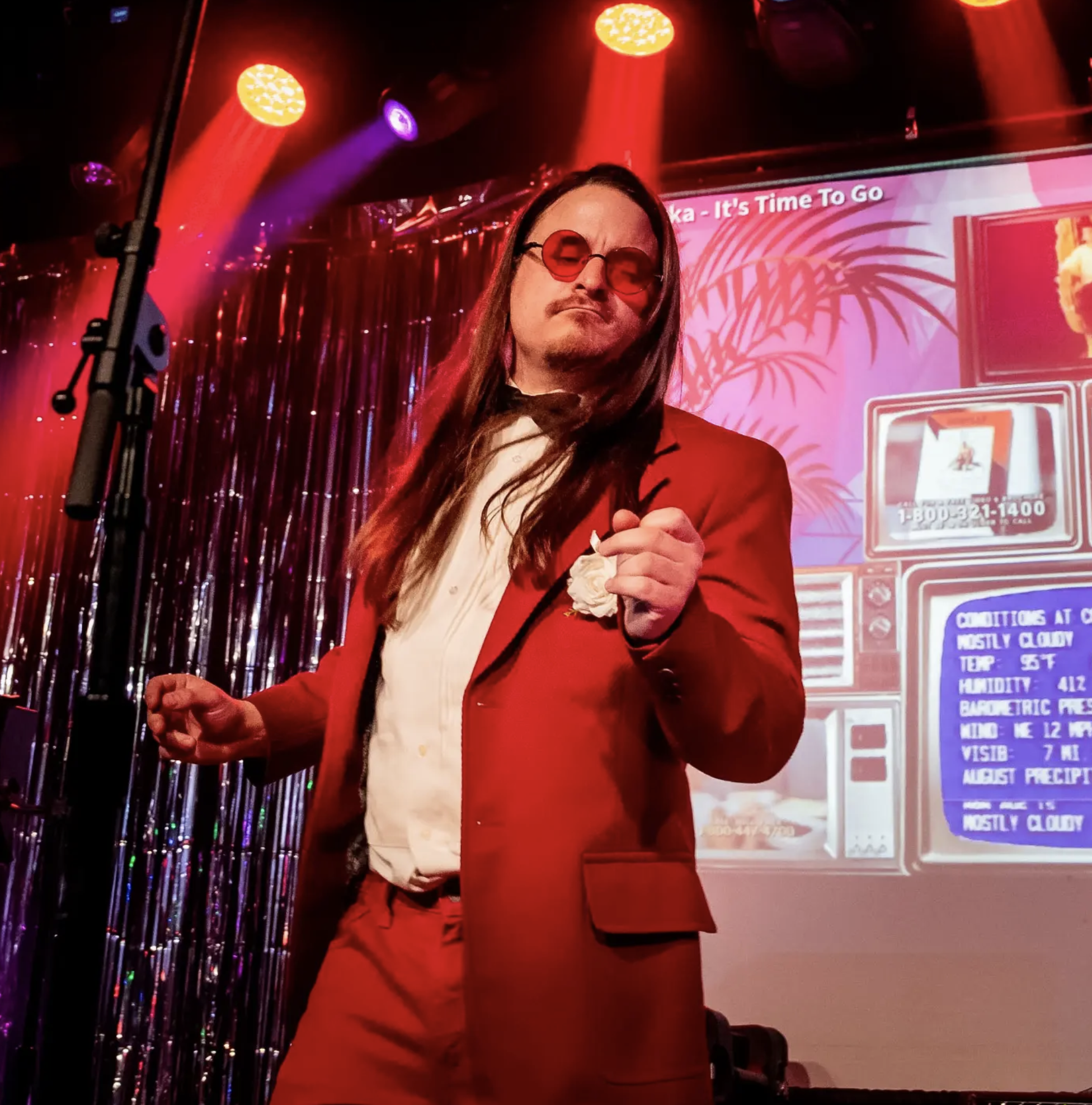 A person with long hair, wearing red tinted glasses and a red jacket, standing in front of colorful stage lights and a pink billboard with tropical design and text, in a vibrant indoor setting.