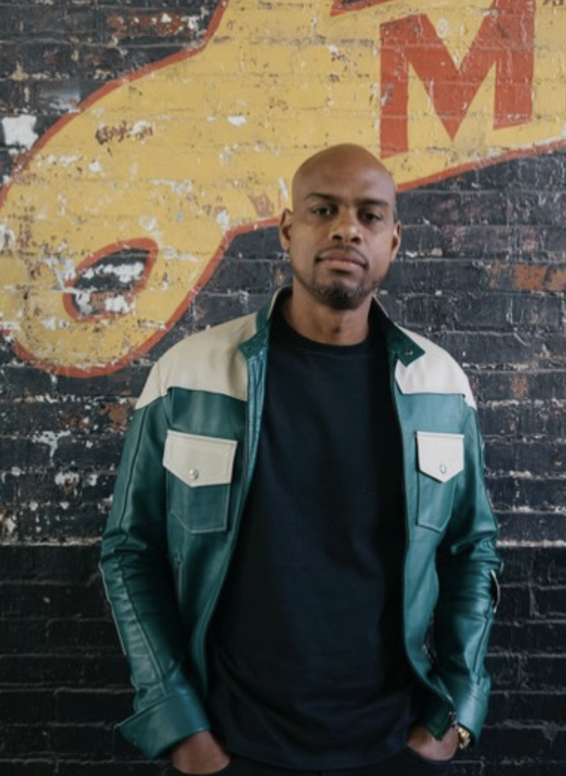 A man standing in front of a brick wall painted with a yellow and red sign. He is wearing a black shirt and a green jacket with beige accents, with his hands in his pockets.