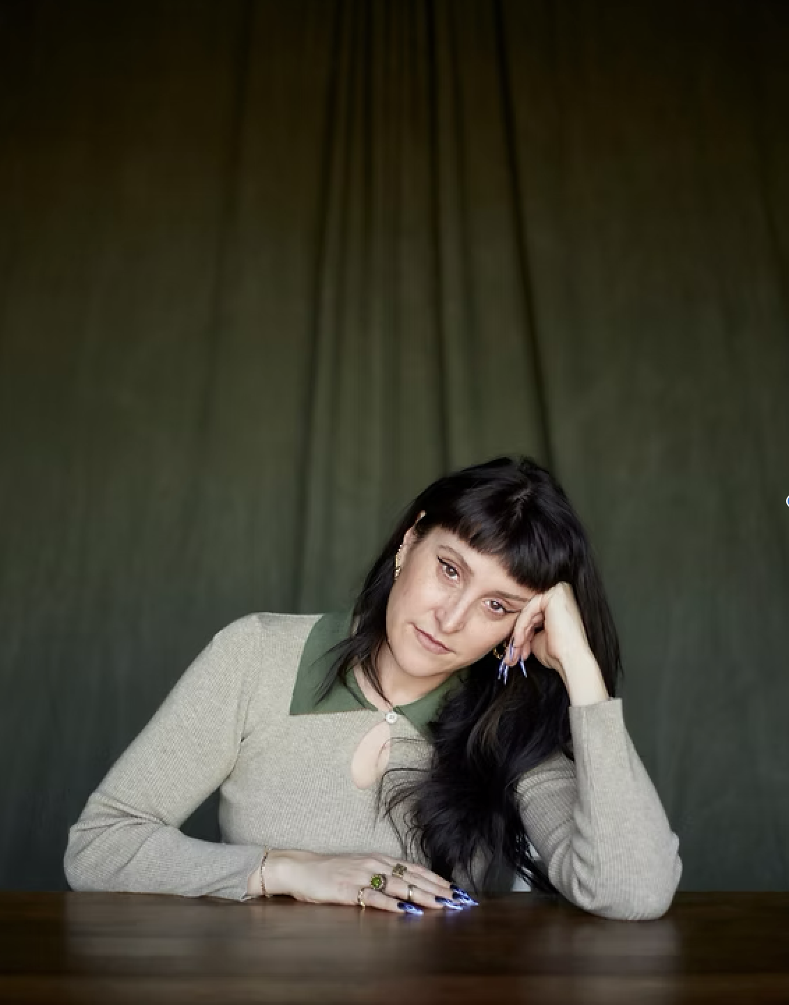 A woman with dark hair and bangs, wearing a beige sweater over a green collared shirt, sitting at a wooden table with a dark green curtain in the background. She is resting her head on her left hand, looking at the camera with a pensive expression.