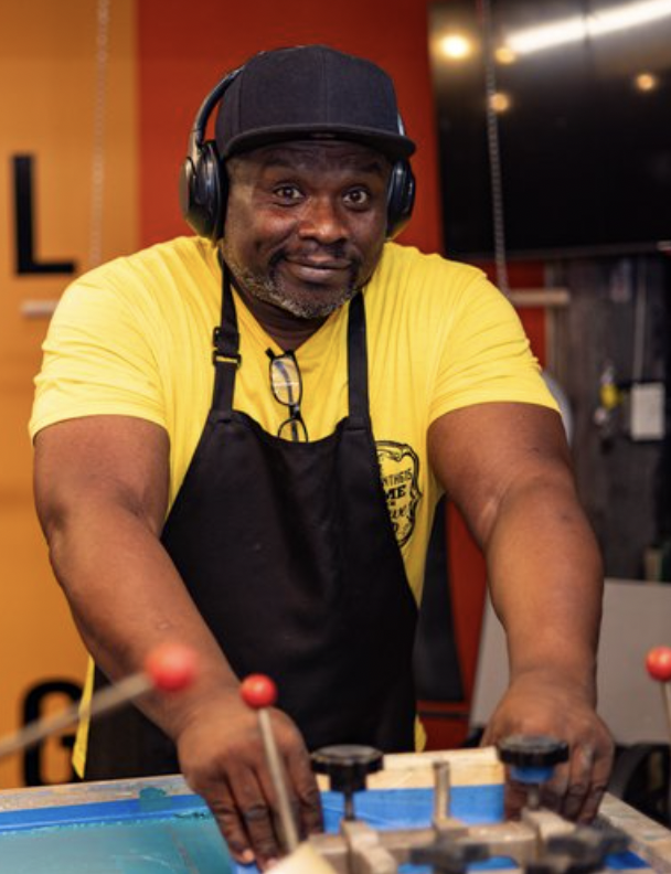 A man in a yellow t-shirt and black apron playing shuffleboard, wearing a black cap and headphones, smiling at the camera in an indoor recreation area.