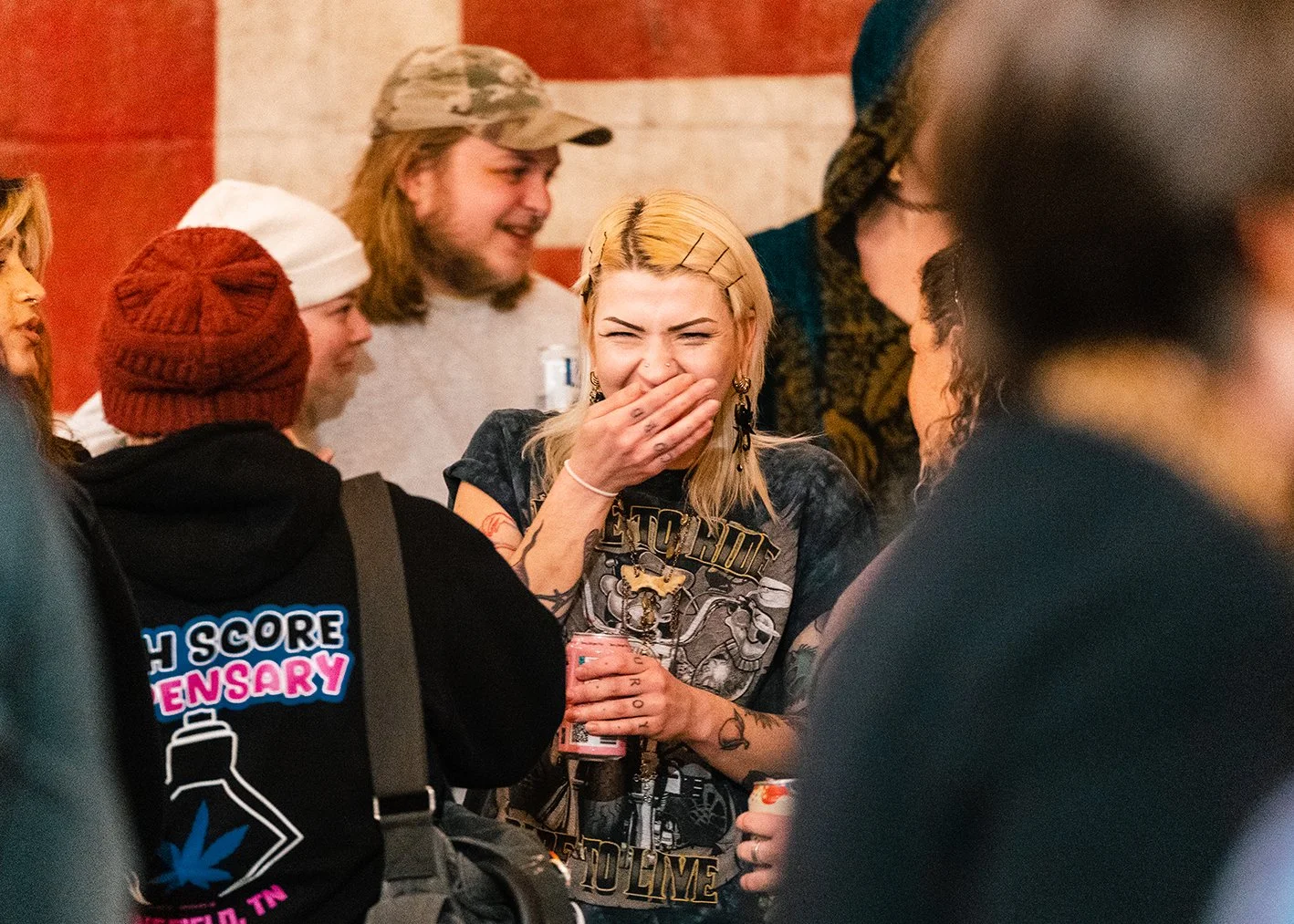 Group of people laughing and talking, with one woman covering her mouth and smiling, holding a drink, in a social setting.