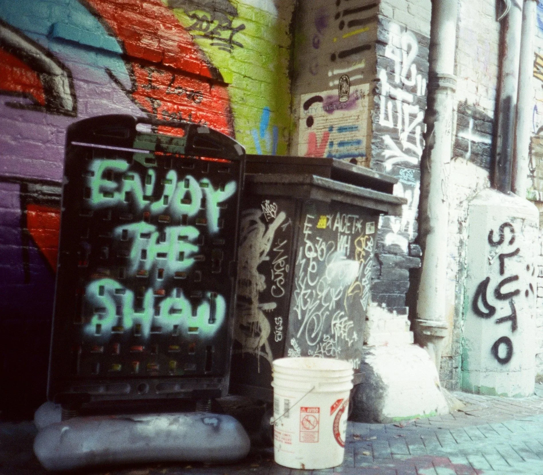 Graffiti-covered brick wall with a black plastic crate, a white bucket of paint, and some pipes. The graffiti includes various words and symbols in multiple colors, with some areas painted in bright colors like purple, green, and yellow.