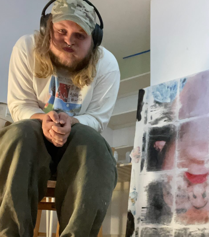 A young man with long blond hair, wearing a camouflage cap, large headphones, a white sweatshirt, and olive green pants, sitting with his hands clasped, in an indoor setting next to a collage or abstract artwork.