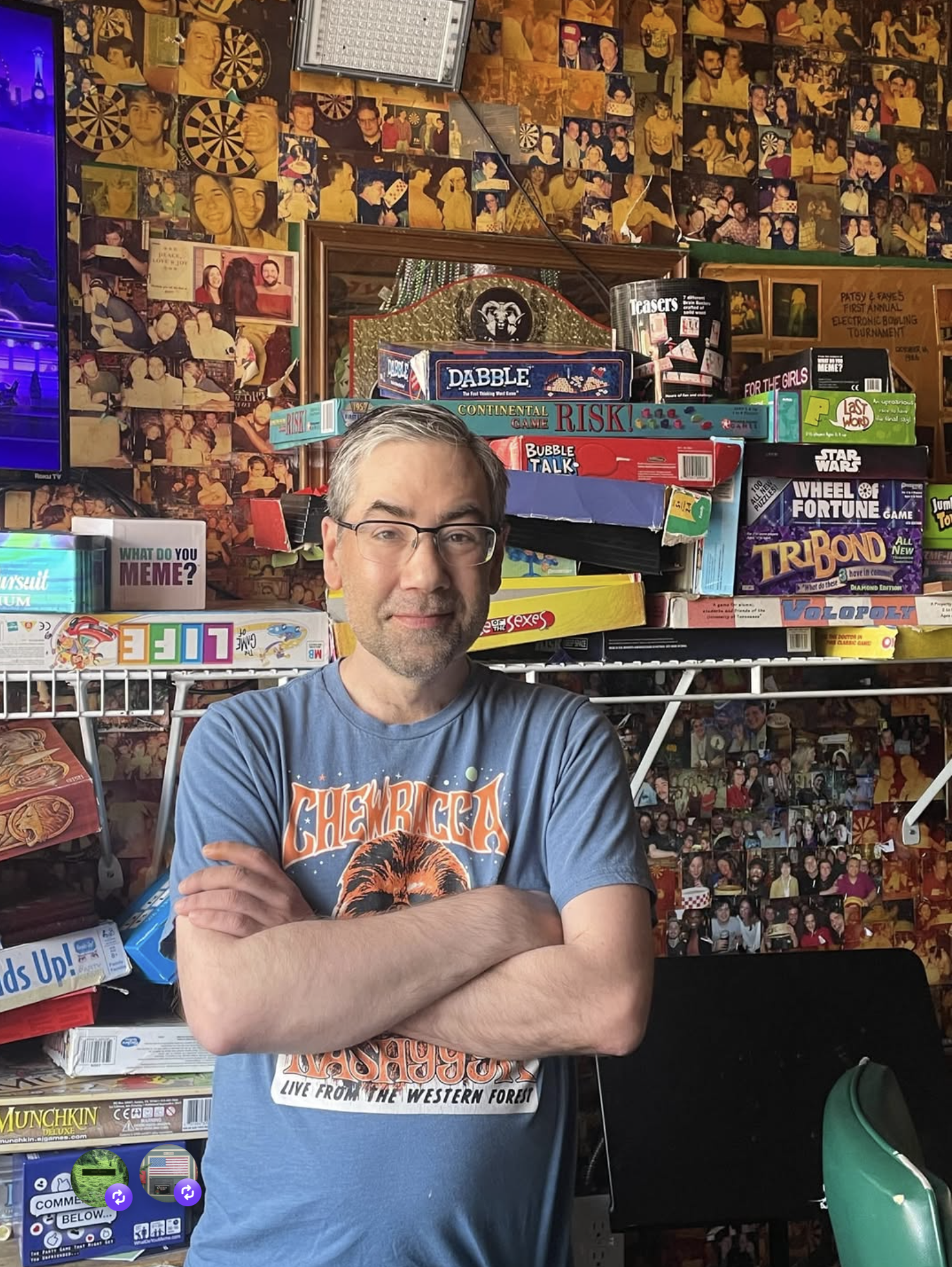 A man wearing glasses and a Chevra shirt standing in front of a wall covered with framed photos and board games, with arms crossed.