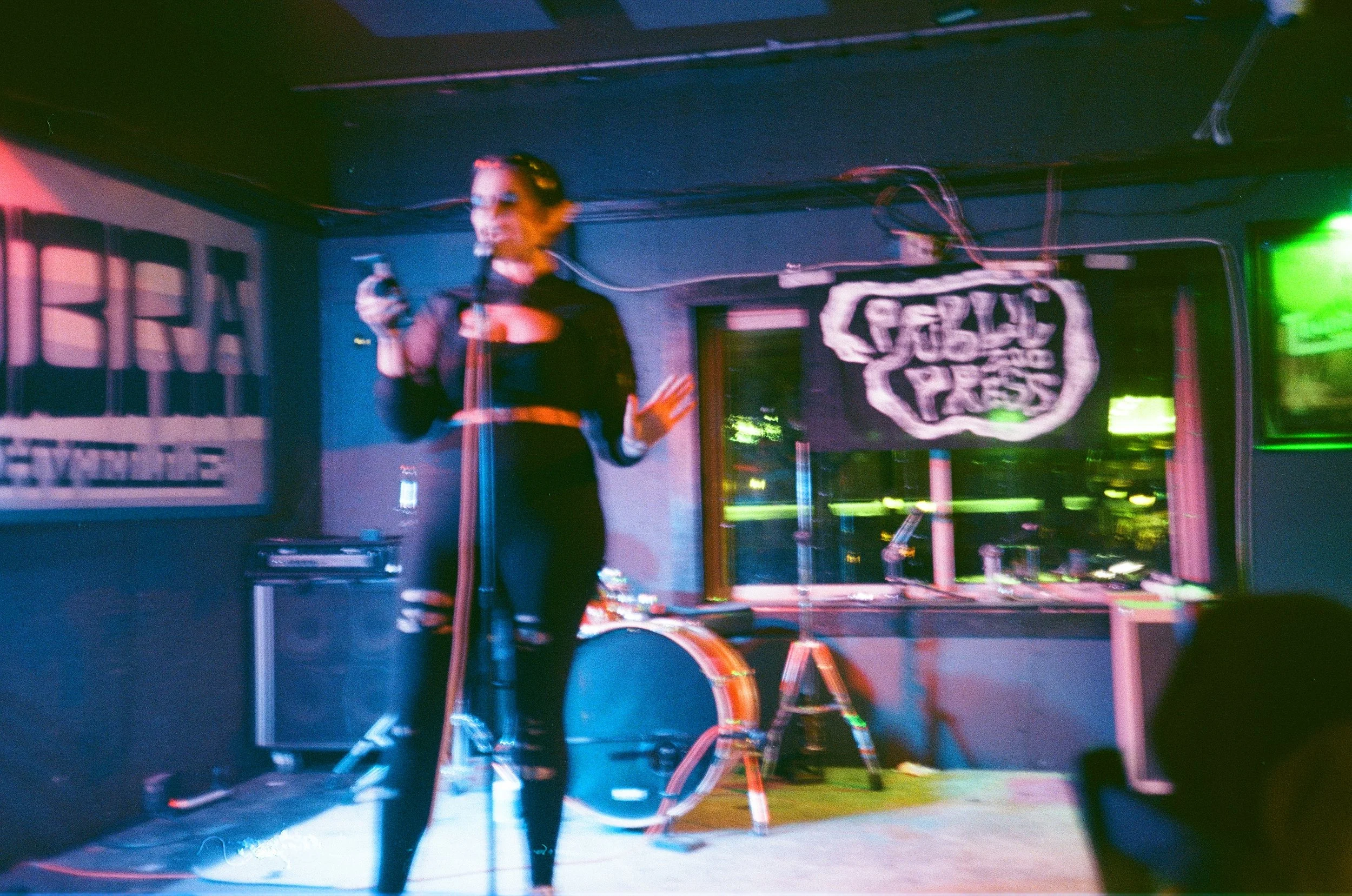A woman speaking into a microphone on stage in a dark venue, with colorful lights and a various equipment and decorations behind her.