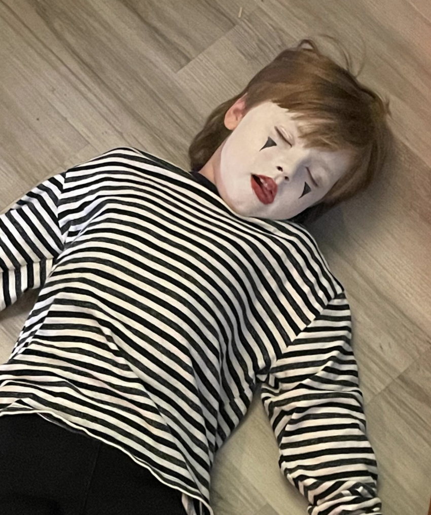 A young boy lying on a wooden floor, dressed as a mime with face paint, a striped shirt, and closed eyes.