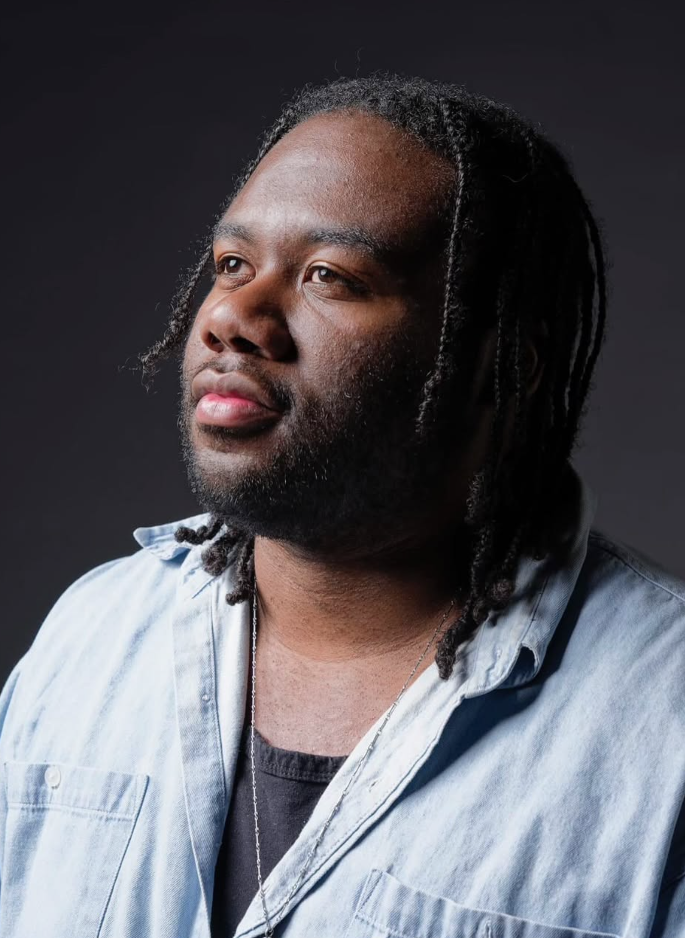 A Black man with dreadlocks and a beard, wearing a light denim shirt over a dark t-shirt, looking slightly to the side against a dark background.