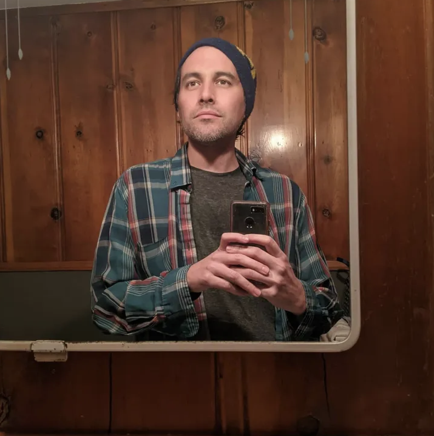 A man taking a selfie in a bathroom mirror, wearing a beanie, a plaid shirt, and a gray t-shirt, with wooden cabinets in the background.