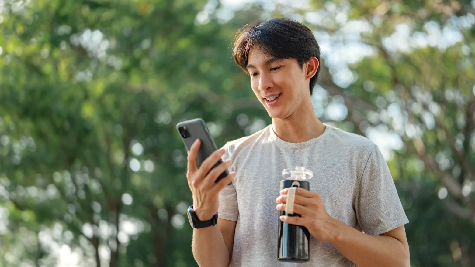 A young man holding a smartphone and a water bottle outdoors in a park with trees in the background.