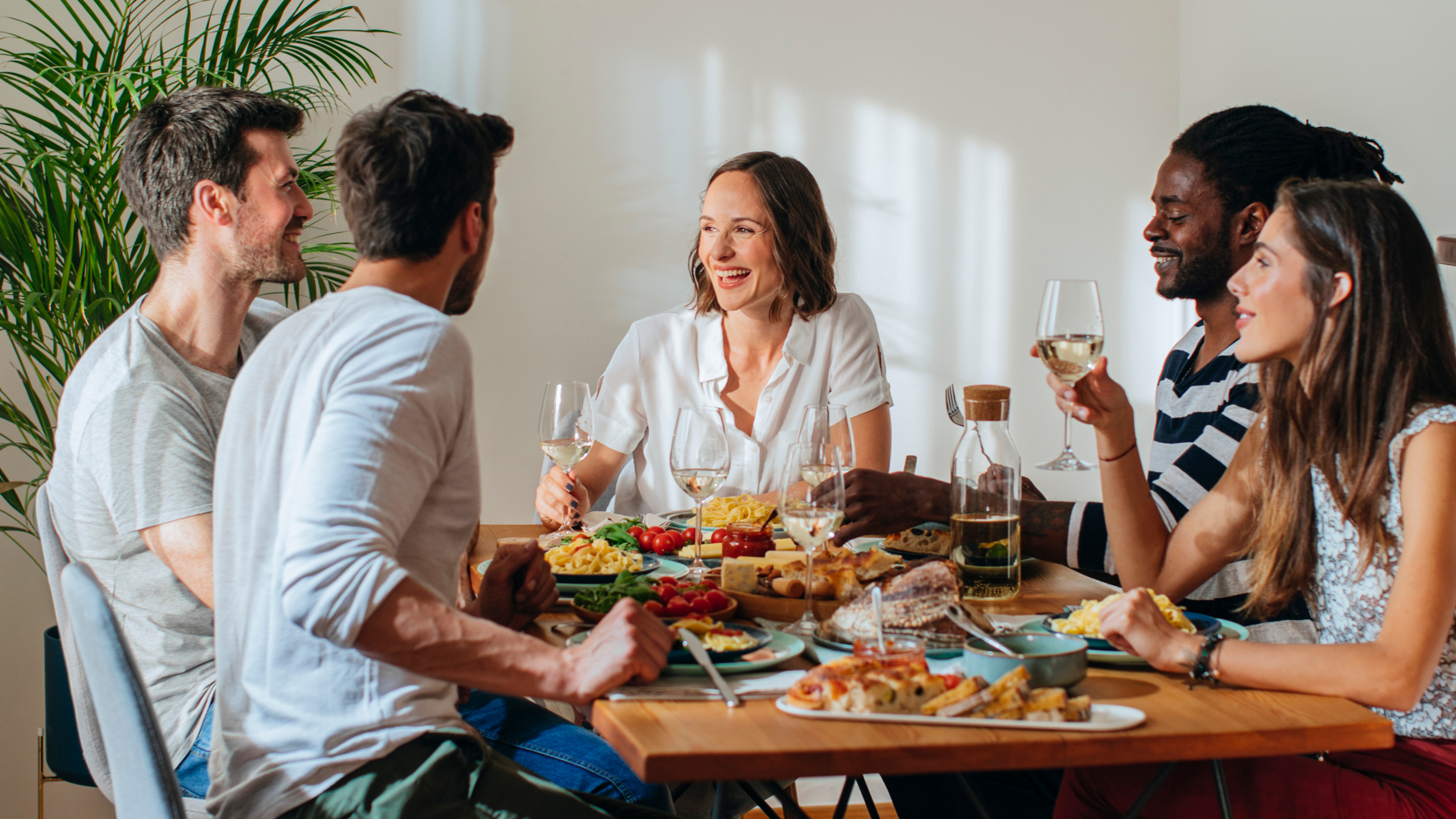 A group of six friends enjoying a meal together at a dinner table, smiling and talking. The table is filled with various dishes including salads, pasta, pizza, and drinks. The setting appears to be a bright, cozy indoor space with a large green plant in the background.