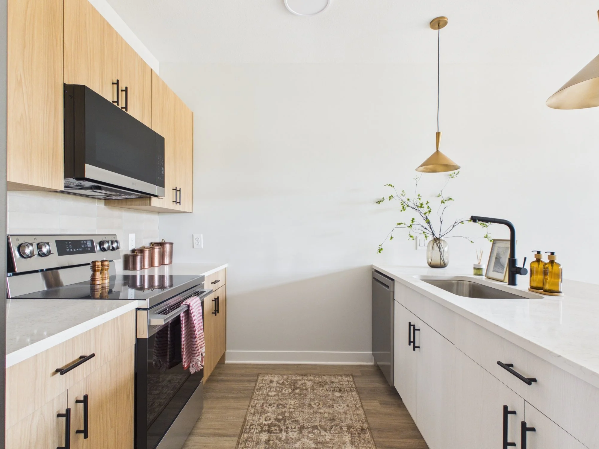 Modern kitchen with light wood and white cabinetry, black hardware, black microwave, stainless steel stove, white countertops, decorative jars, potted plant, framed photo, and gold pendant lights.