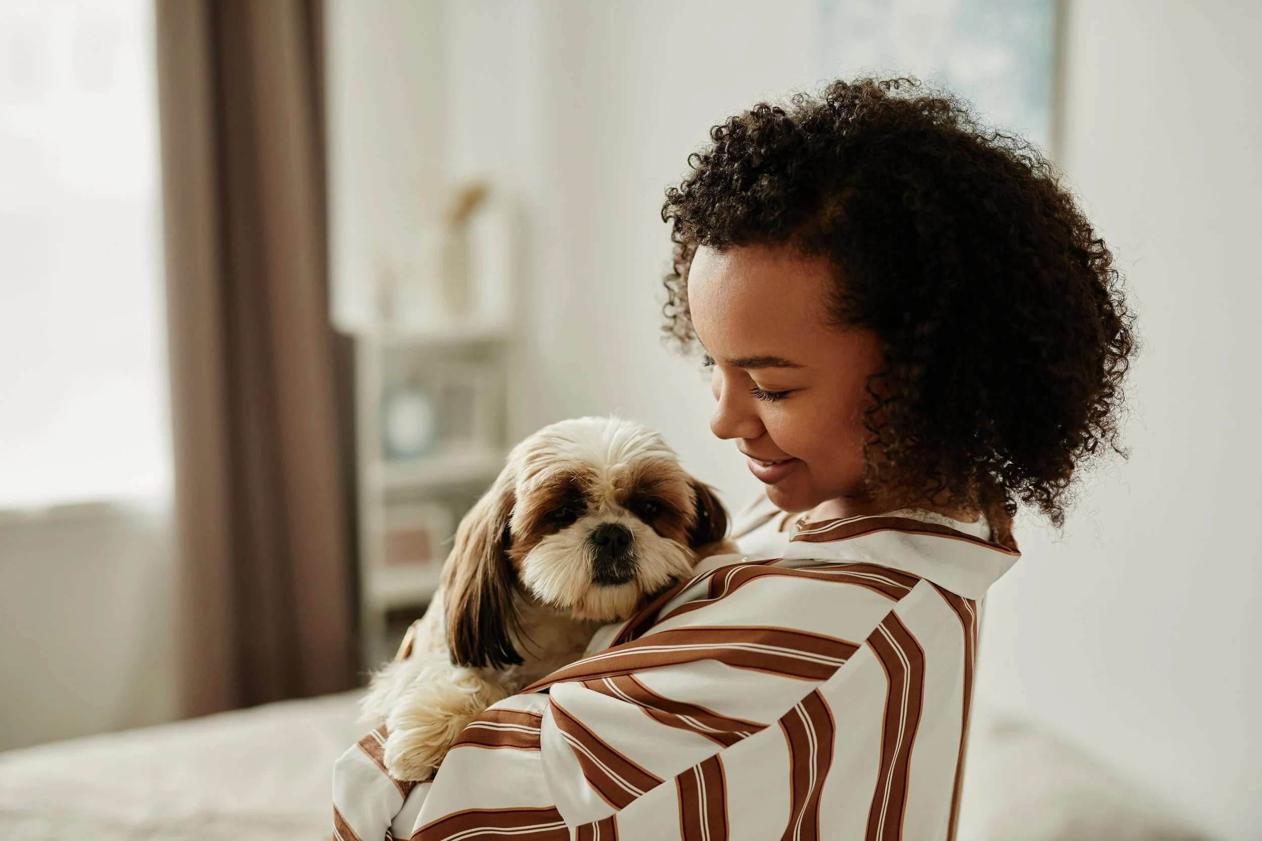 A woman with curly hair smiling while holding a small fluffy dog with closed eyes.