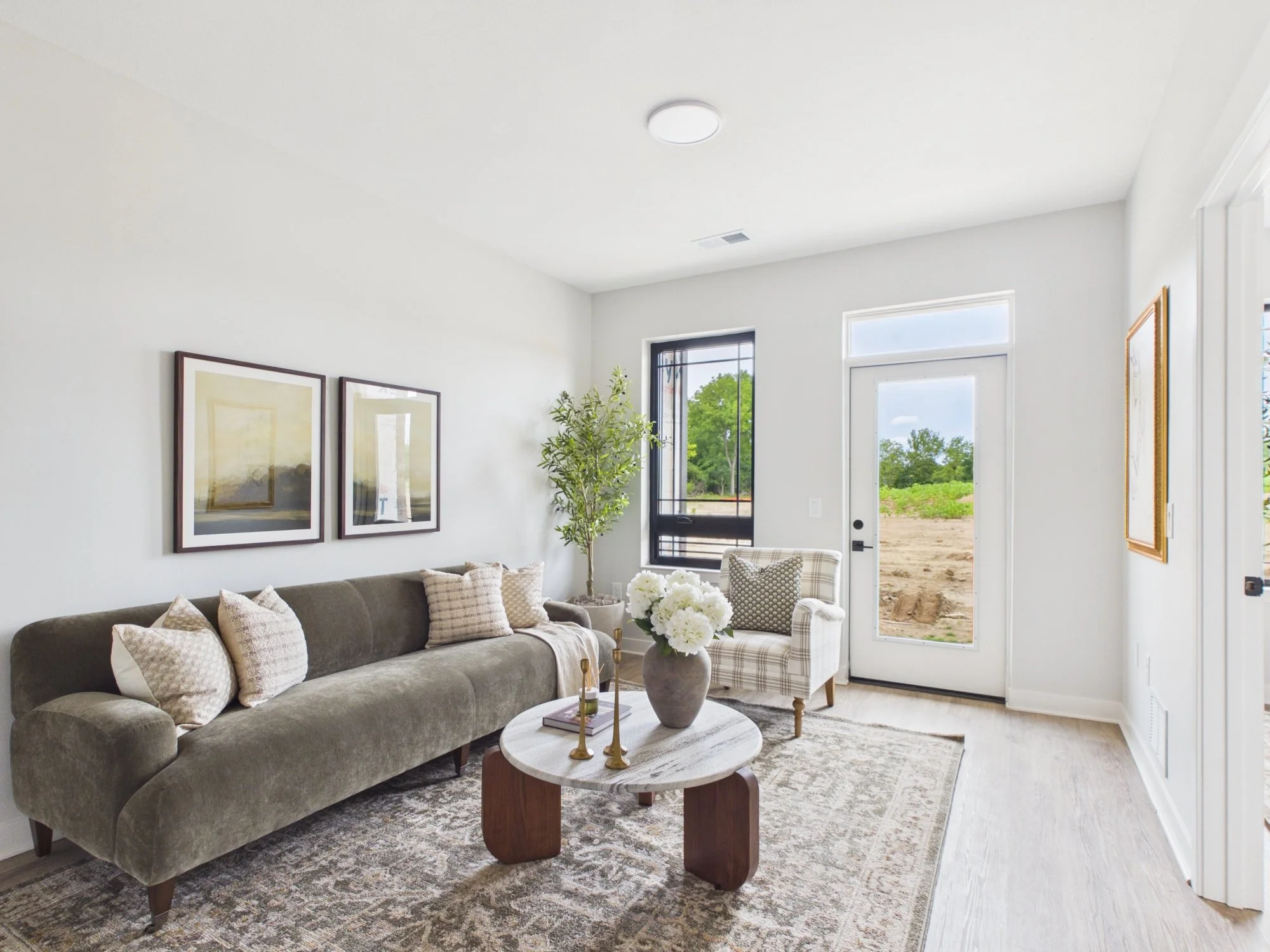 Modern living room with gray sofa, patterned pillows, white round coffee table with decorative candles and flowers, framed artwork, green plant, and large windows showing outdoor landscape.