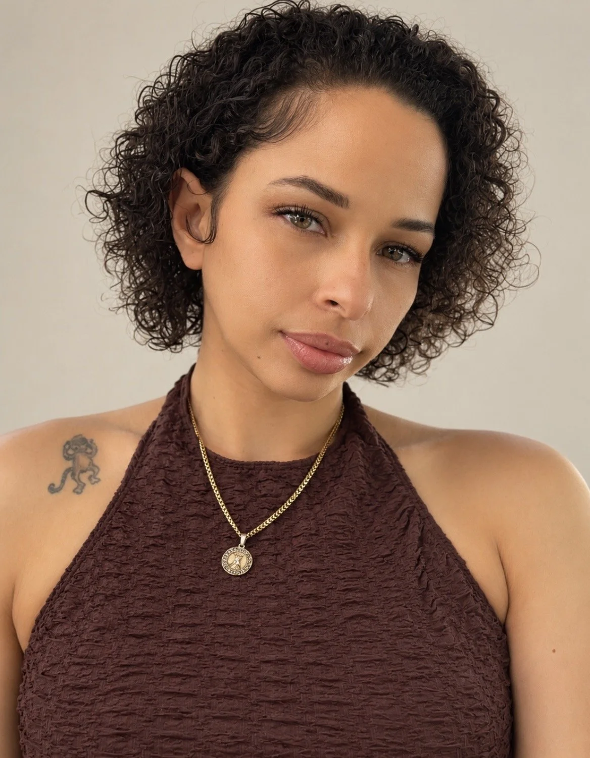 Close-up portrait of a young woman with curly dark hair, light skin, and grey eyes. She is wearing a sleeveless maroon textured top, a gold chain necklace with a circular pendant, and has a small lion tattoo on her left shoulder. The background is neutral.