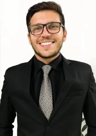 Homem sorridente vestindo terno preto, camisa preta, gravata e óculos, posando contra fundo branco.